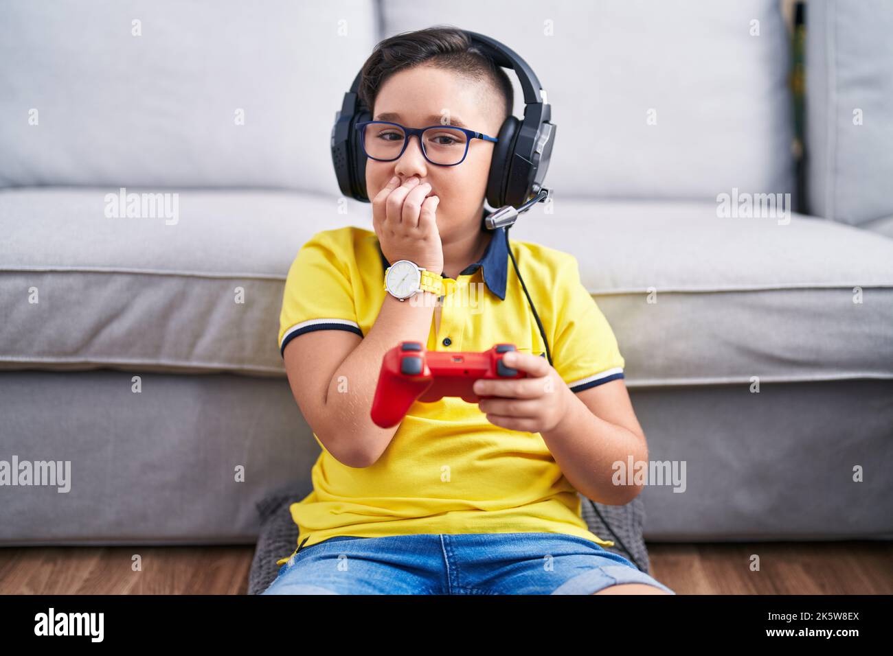 Young hispanic kid playing video game holding controller wearing headphones laughing and embarrassed giggle covering mouth with hands, gossip and scan Stock Photo