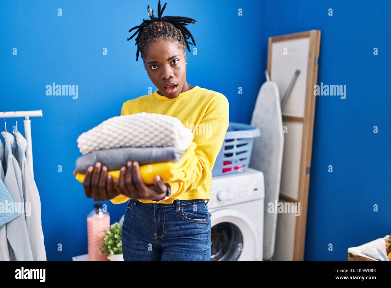 Beautiful black woman holding clean laundry in shock face, looking