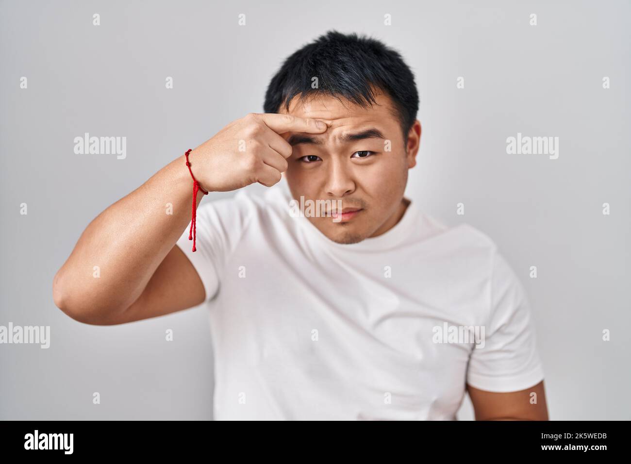Young chinese man standing over white background pointing unhappy to pimple on forehead, ugly ...