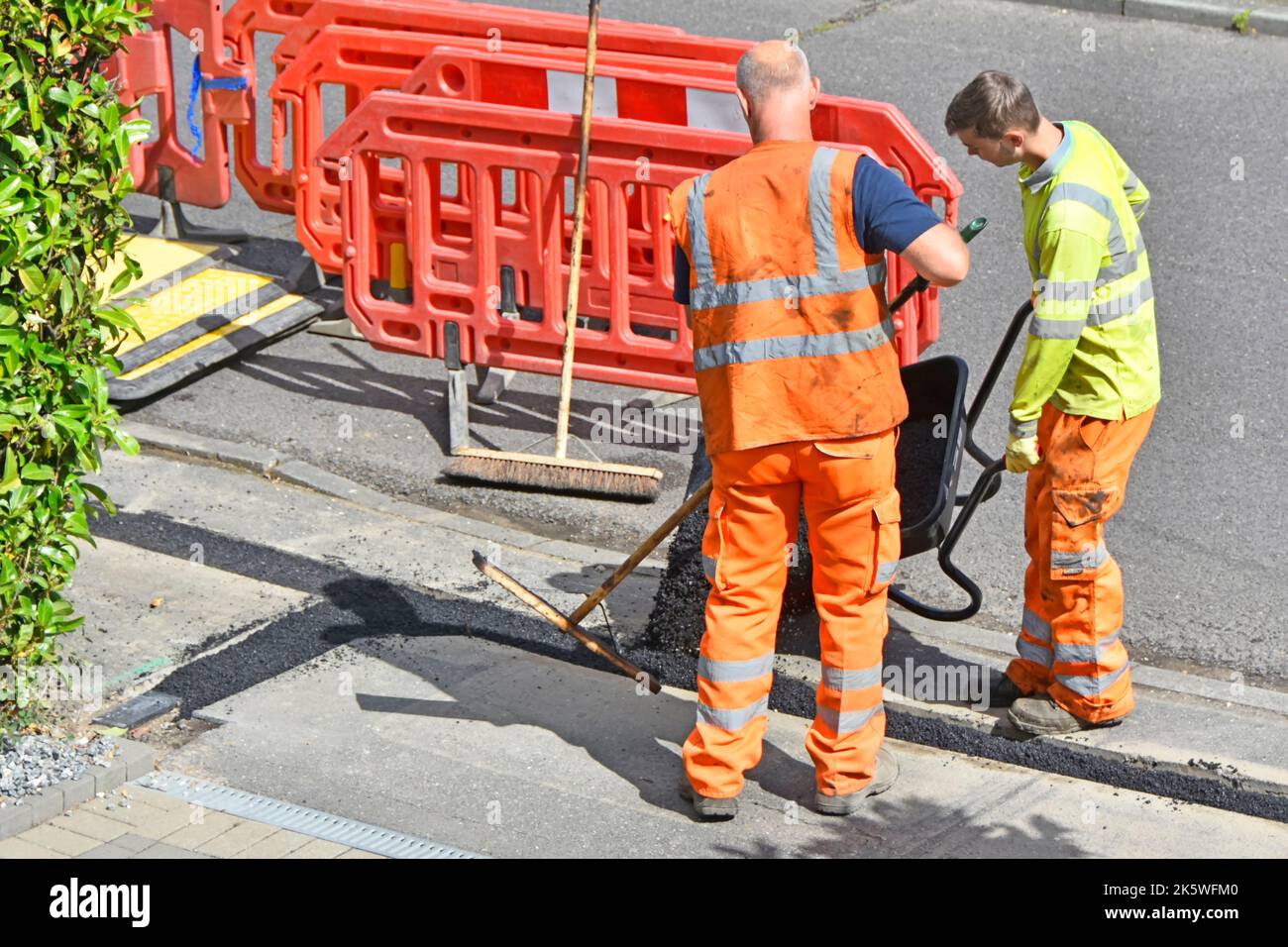 Workmen tip tarmac type paving material from wheelbarrow to reinstate narrow pavement trench after underground fibre optic broadband cable laying UK Stock Photo