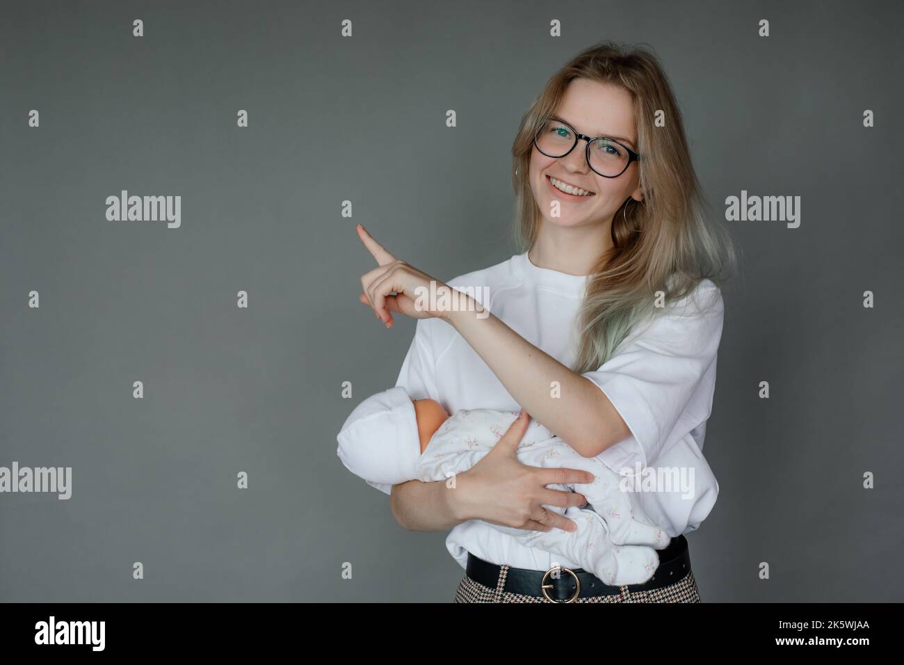 Portrait of young cheerful woman holding baby doll in white clothes, pointing with index finger on grey background. Stock Photo