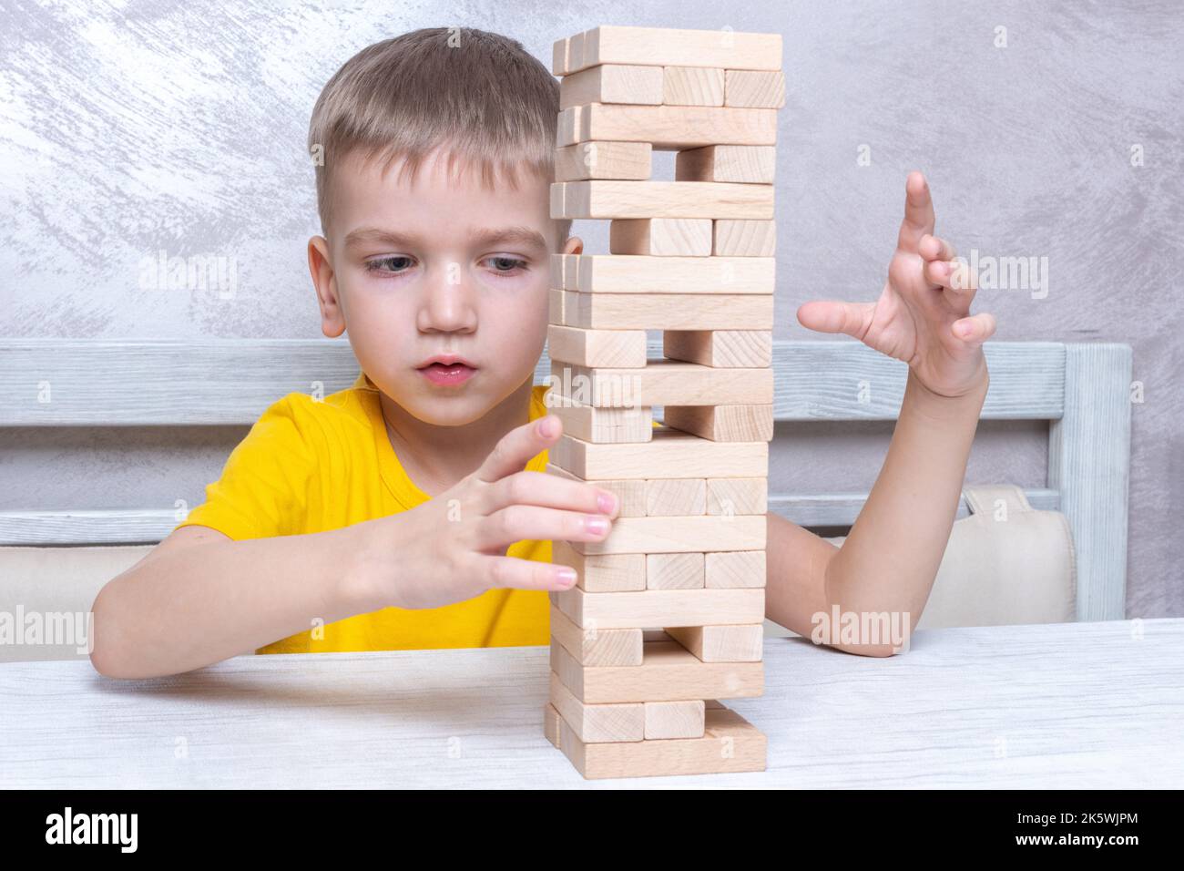 Interested happy little blond boy playing board game taking bricks from wooden tower keeping balance. Board game Tower of wooden blocks. Activity for Stock Photo