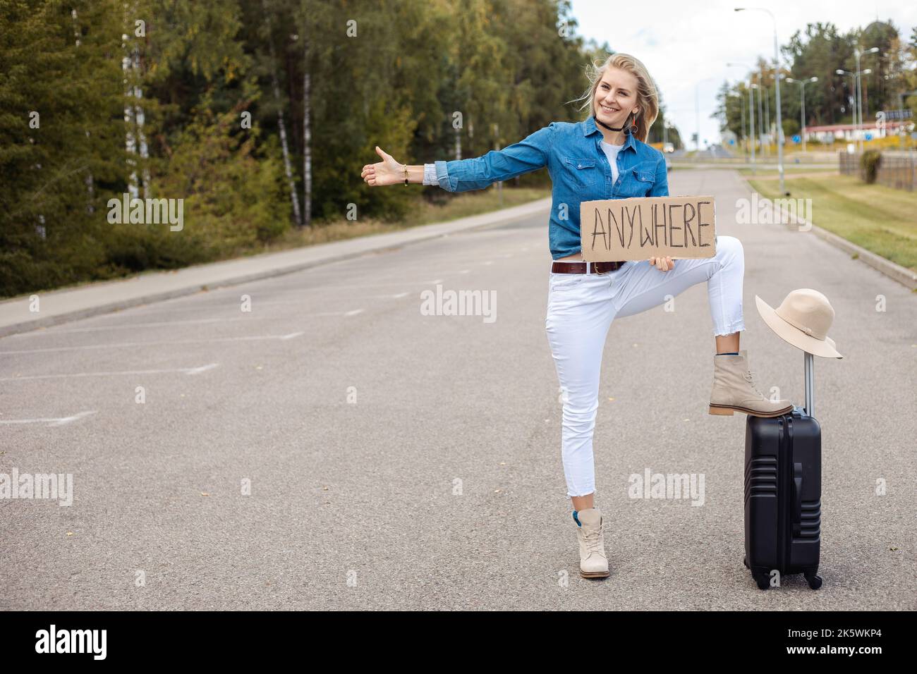 Young woman standing on road with leg on black suitcase, holding cardboard with inscription anywhere, thumbing ride. Stock Photo