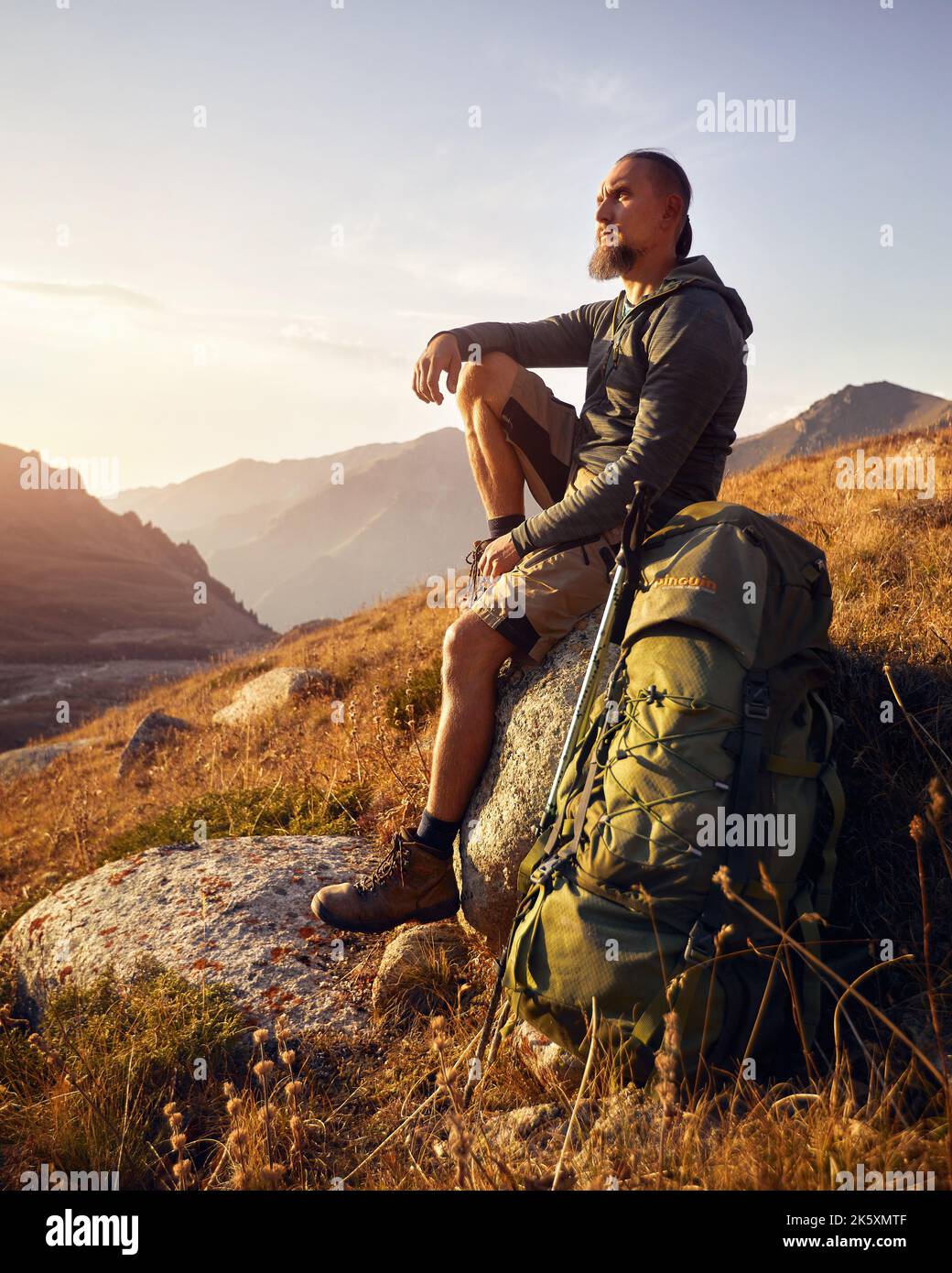 Portrait of Bearded man hiker tourist with big backpack and trekking poles sitting  on the rock against beautiful sunset in the mountain valley. Outdo Stock Photo