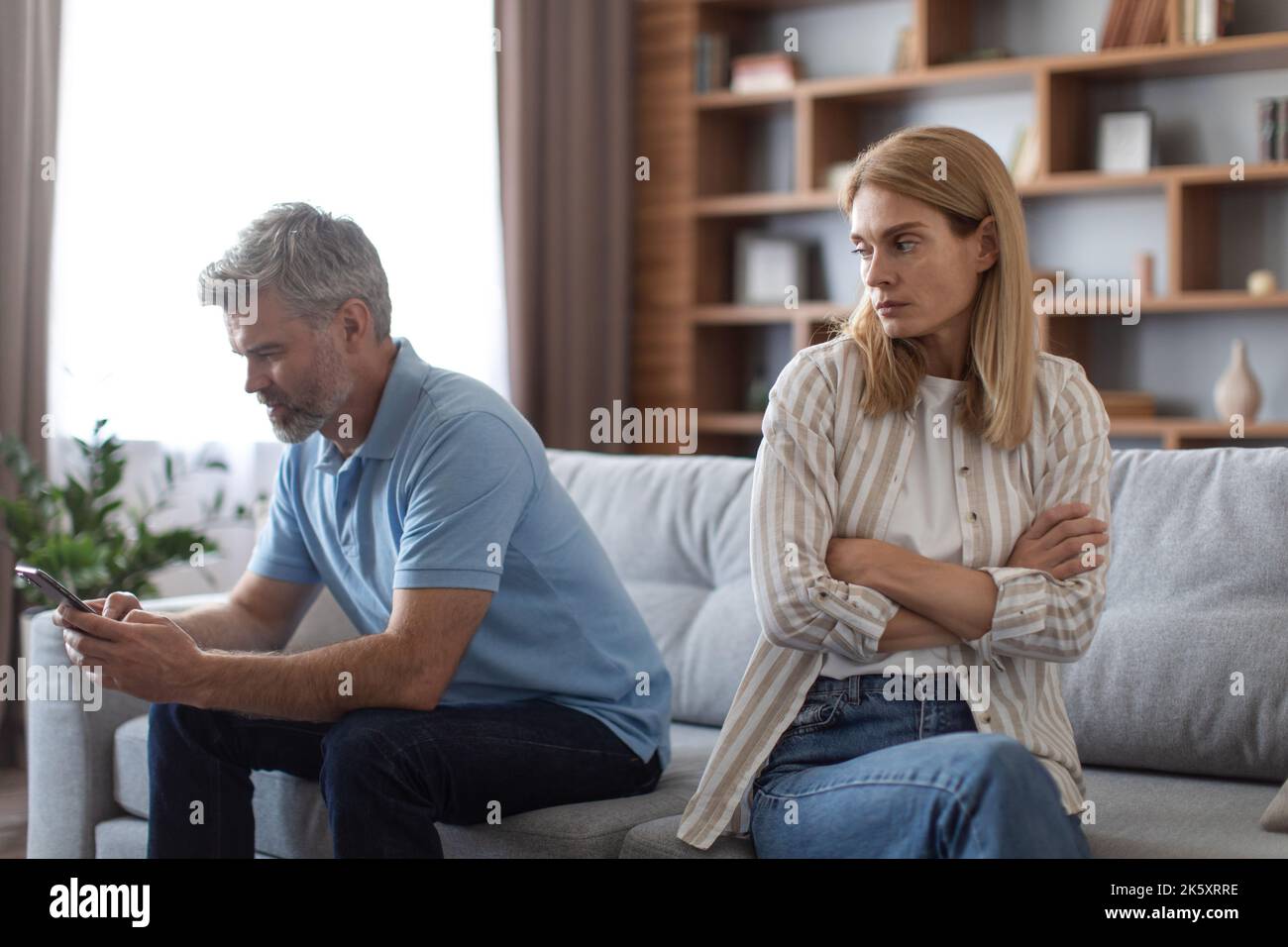 Sad middle aged european wife is jealous of her husband with smartphone in living room interior, copy space Stock Photo