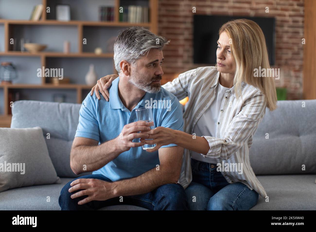 Sad middle aged european wife gives water and calms depressed unhappy man in living room interior Stock Photo