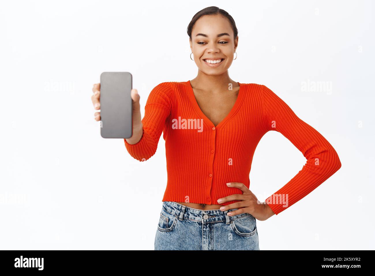 Portrait of 30 years old woman showing smartphone screen, demonstrates app interface, webstore, stands with mobile phone against white background Stock Photo