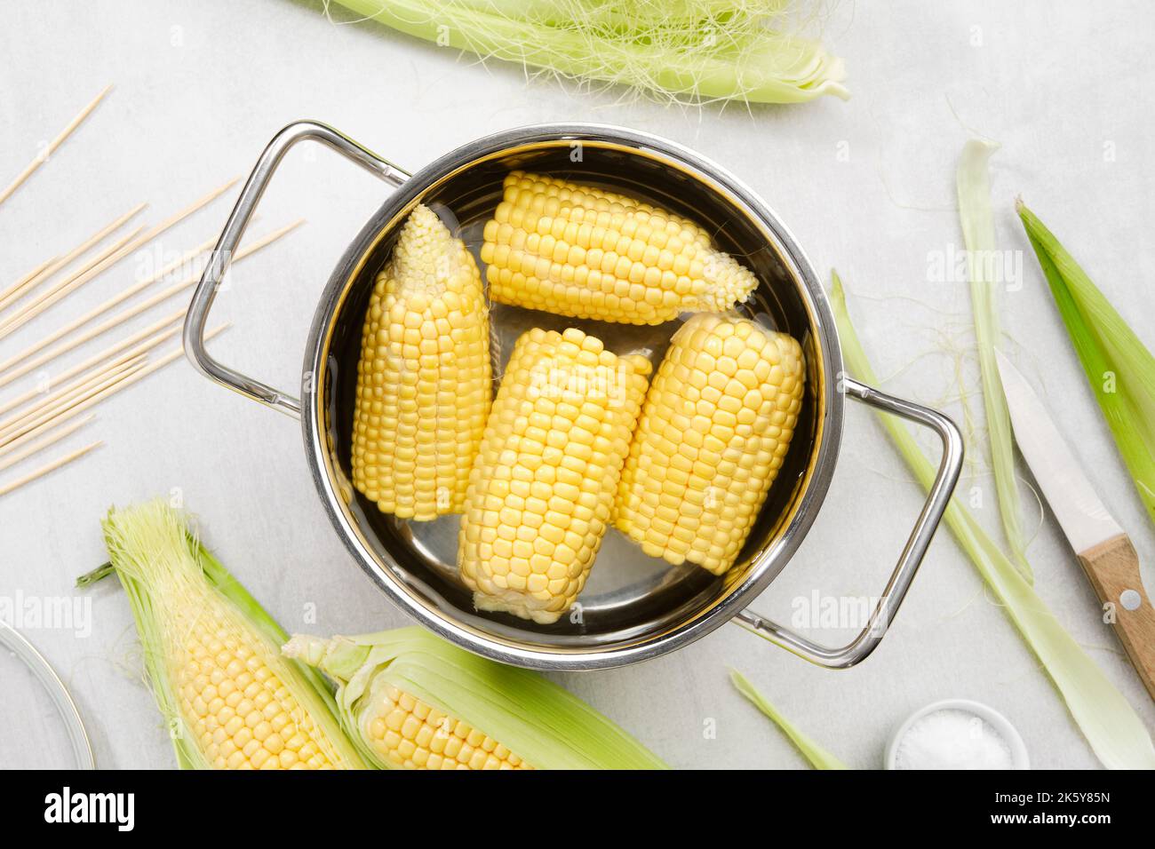Cooking pot of yellow sweet corn cobs prepared for cooking, raw corncobs, wooden skewers, knife and salt on kitchen table, top view. Stock Photo