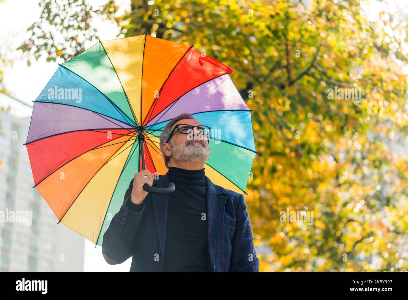 Volatile autumn weather. Lazy Sunday walk in nearby park. Medium closeup outdoor shot of elegant caucasian bearded man with black glasses holding big vivid rainbow umbrella and looking at the sky. Blurred background with tree that changed leaf color. High quality photo Stock Photo