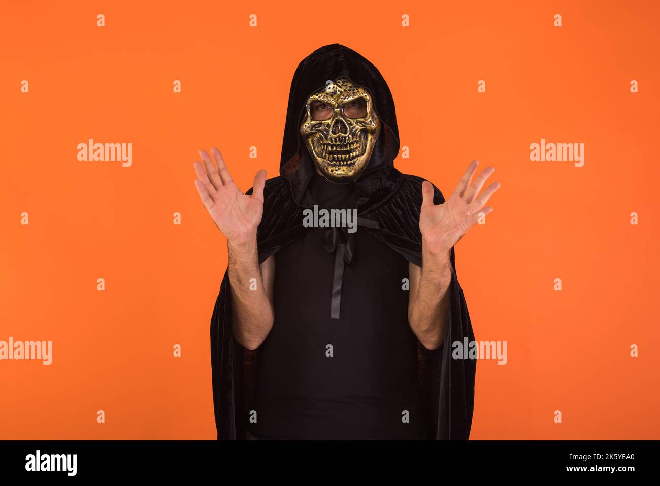 Person with skull mask and black hooded cape, waving with hands, celebrating Halloween, on orange background. Celebration concept, All Souls' Day and Stock Photo