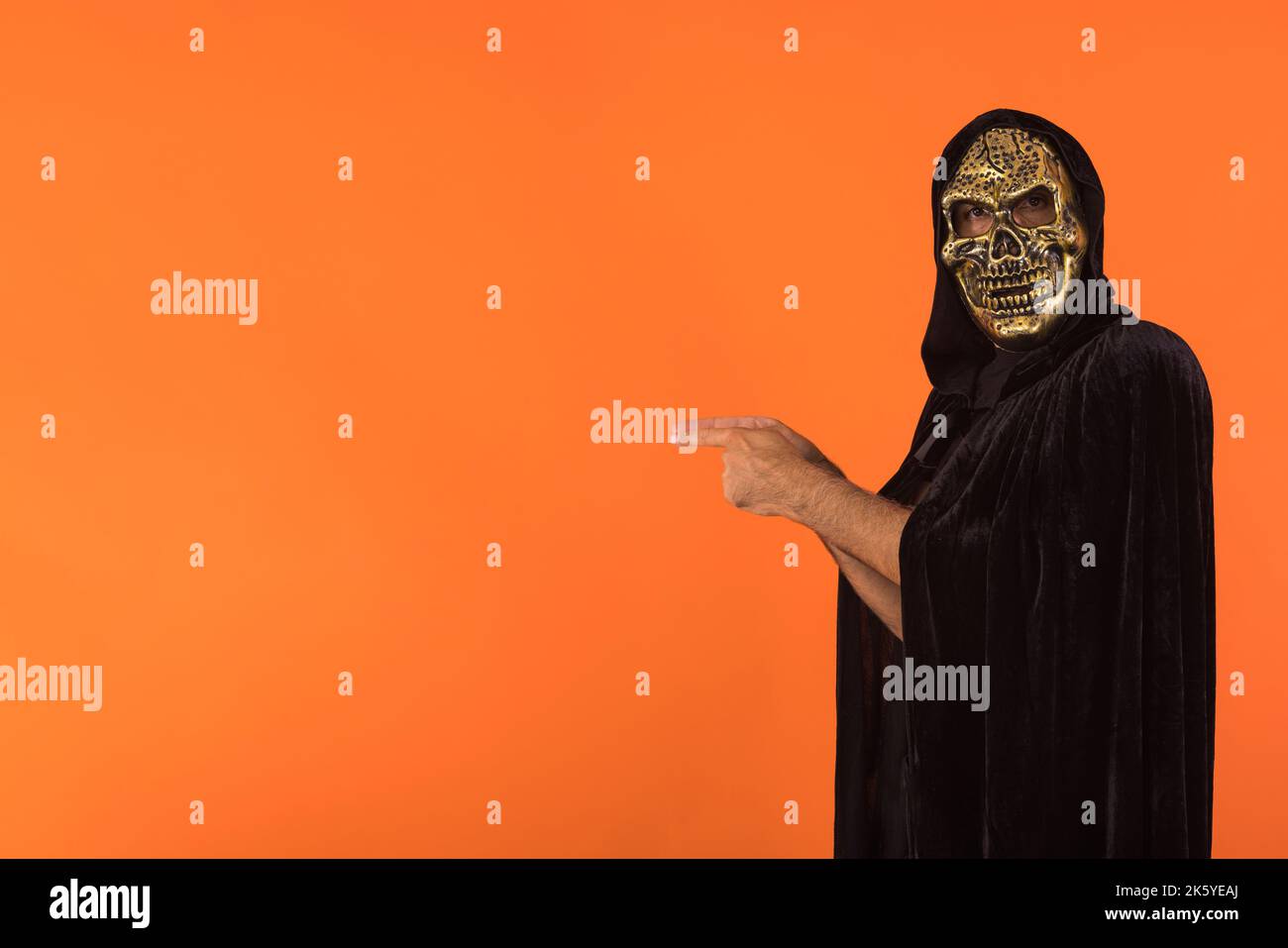 Person with skull mask and black hooded cape, pointing fingers to the side, celebrating Halloween, on orange background. Celebration concept, All Soul Stock Photo