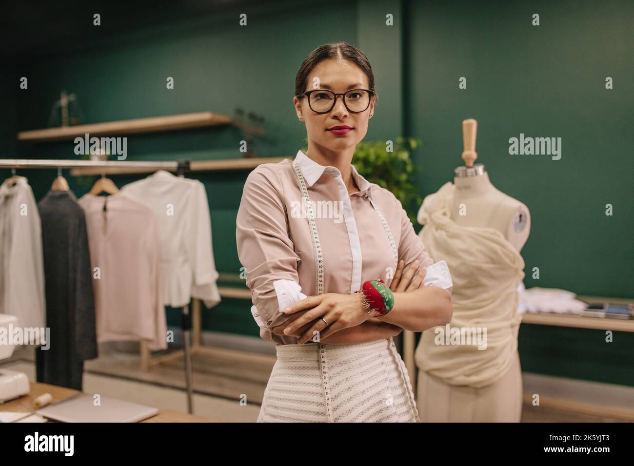 Successful female designer with measuring tape around her neck standing in her workshop. Woman standing with arms crossed in fashion studio. Stock Photo
