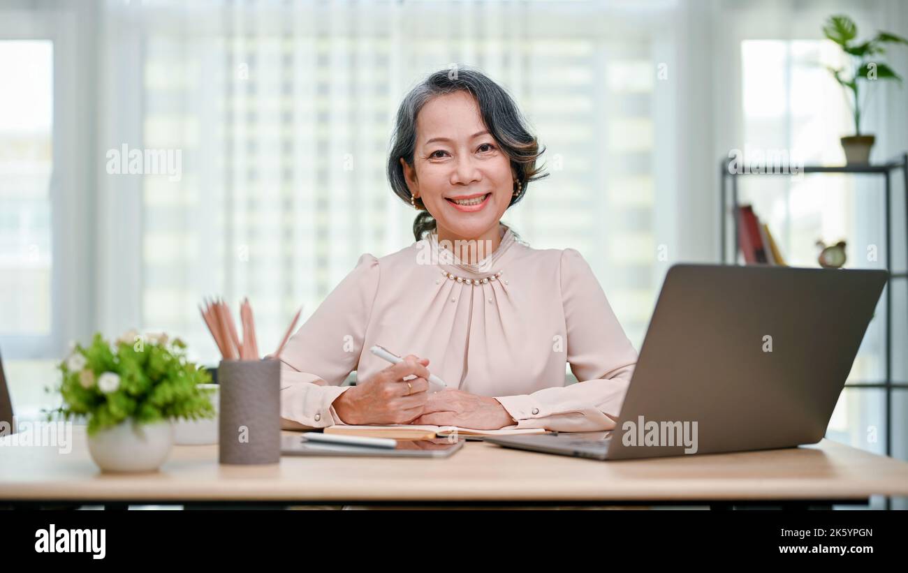 Happy and charming 60s aged-asian woman or businesswoman sitting at her office desk, smiling and looking at the camera. Stock Photo
