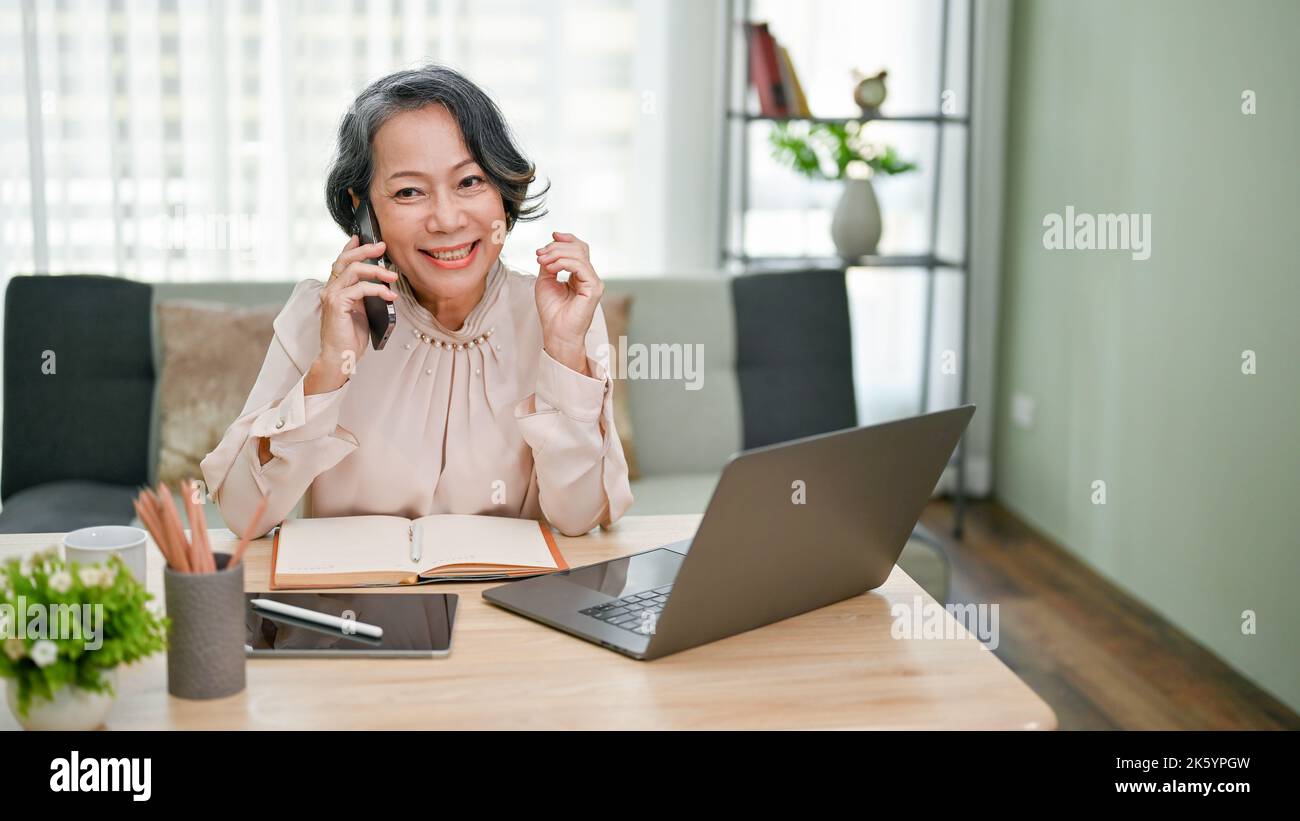 Charming and cheerful senior asian businesswoman talking on the phone with her business supplier while working at her desk in the office. Stock Photo