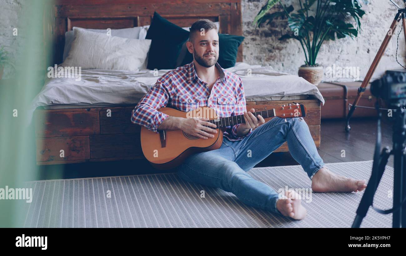 Handsome man blogger and guitarist is recording video lesson about guitar in bedroom using camera. Bearded guy is greeting his followers, talking and showing guitar parts. Stock Photo