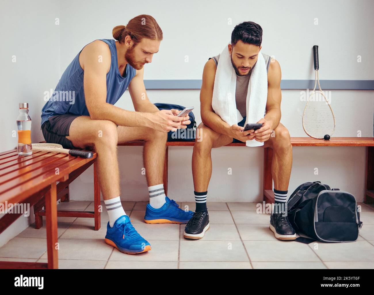 Two players using their cellphones together. Young squash players reading text messages on their smartphones together. Friends relaxing in a gym Stock Photo