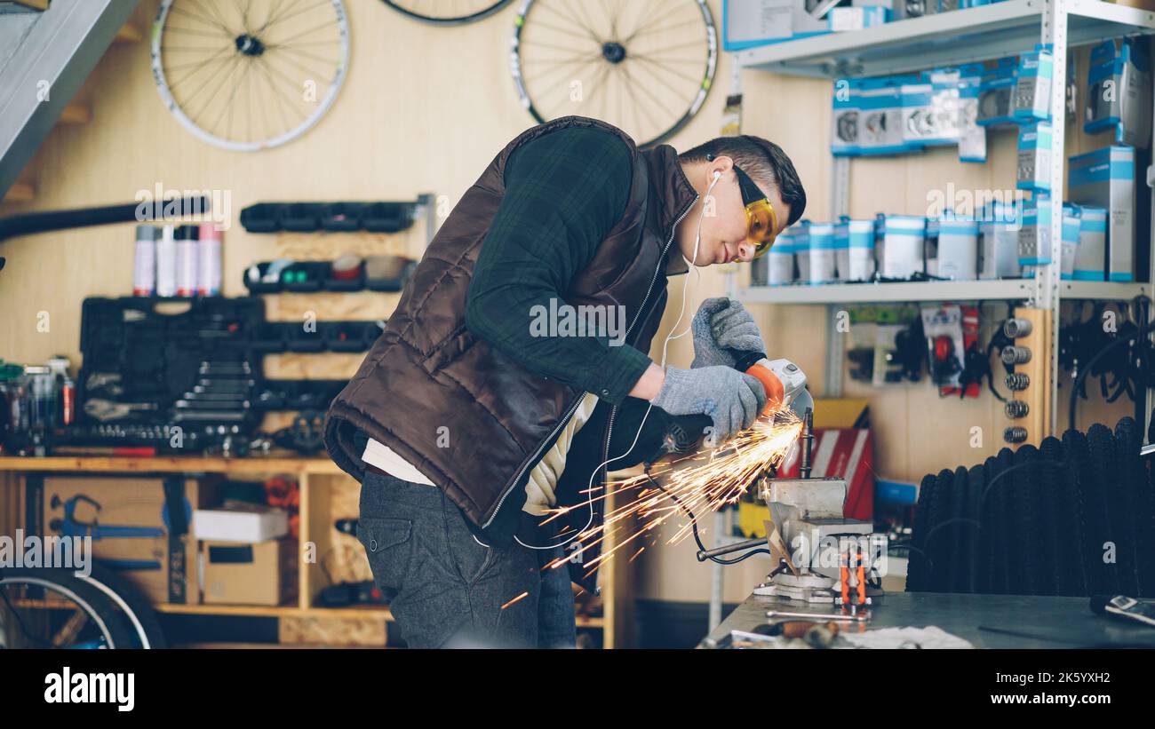 Experienced maintenance man is using electric circular saw while working in his small workplace. Young man is wearing protective glasses and gloves and is listening to music with earphones. Stock Photo
