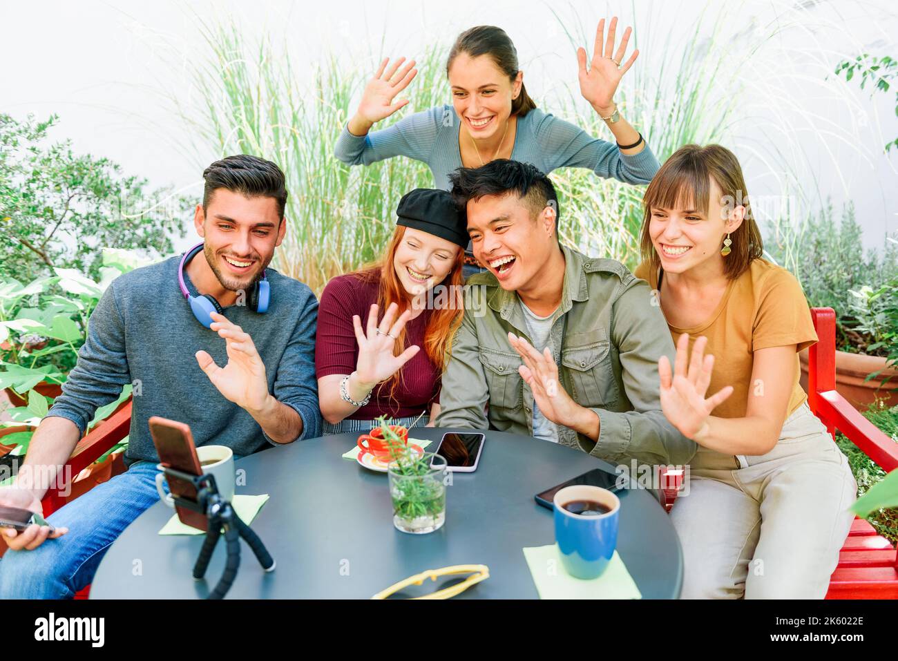 From above happy multiracial men and women smiling and waving hands while sitting at cafe table and making video call in garden Stock Photo