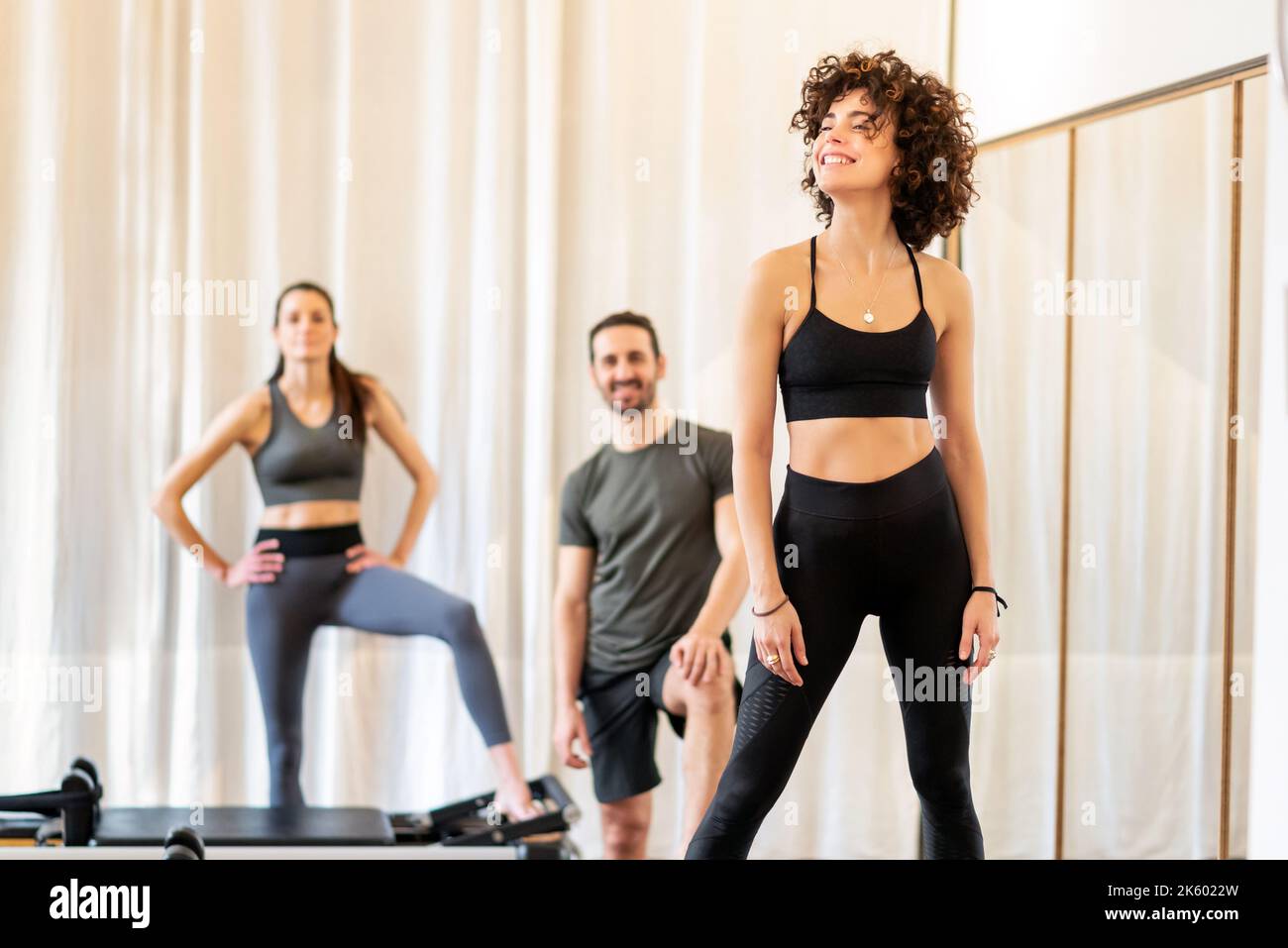 Smiling fit woman with curly hair standing in fitness studio with cheerful friends near reformer while resting during pilates session Stock Photo