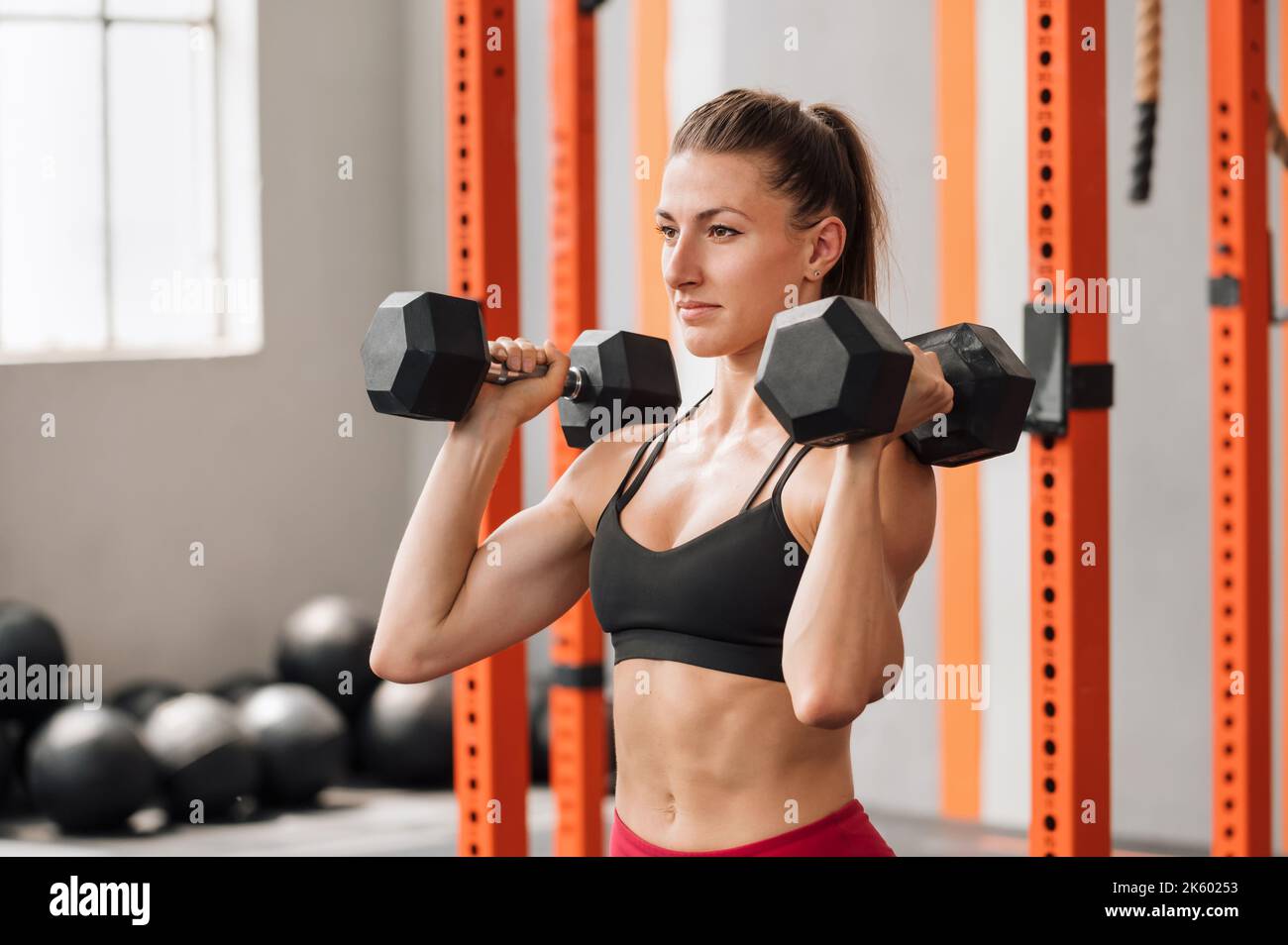 Strong female athlete in sports bra doing dumbbell press exercise while working out in modern gym Stock Photo
