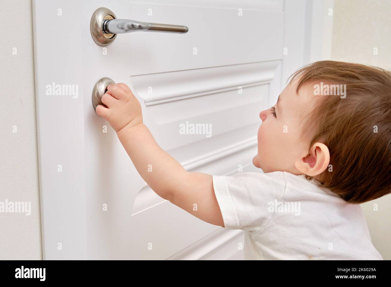 Toddler baby opens the lock, child hand closeup. White wooden door
