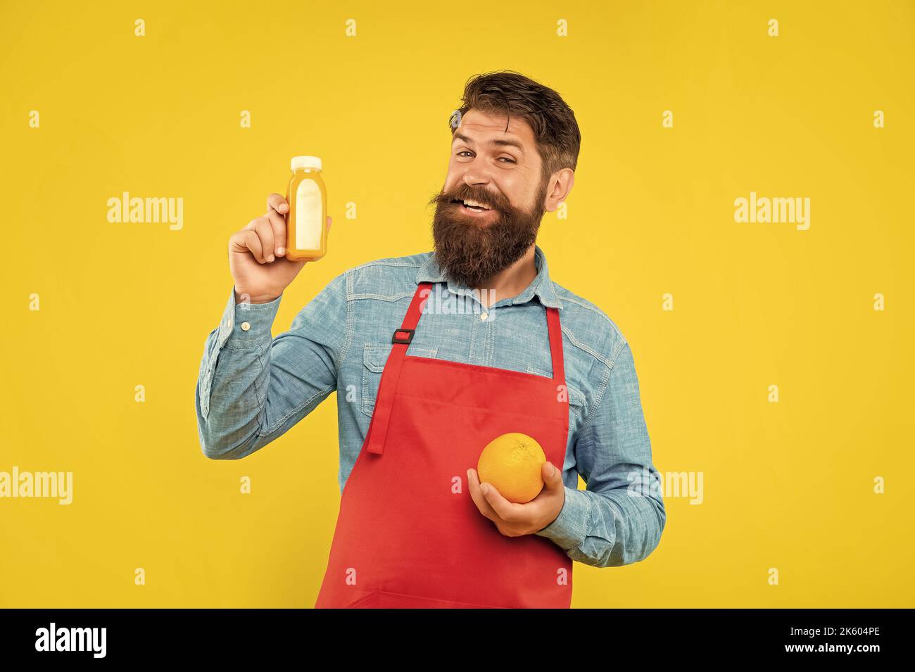 Happy man in apron holding orange and juice bottle yellow background, barkeeper Stock Photo