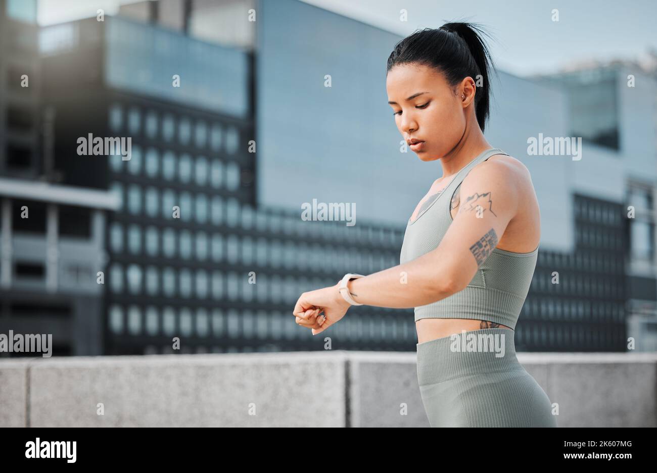 Young hispanic fit female athlete with tattoos looking serious while checking the time on her watch to track her progress while taking a break from Stock Photo