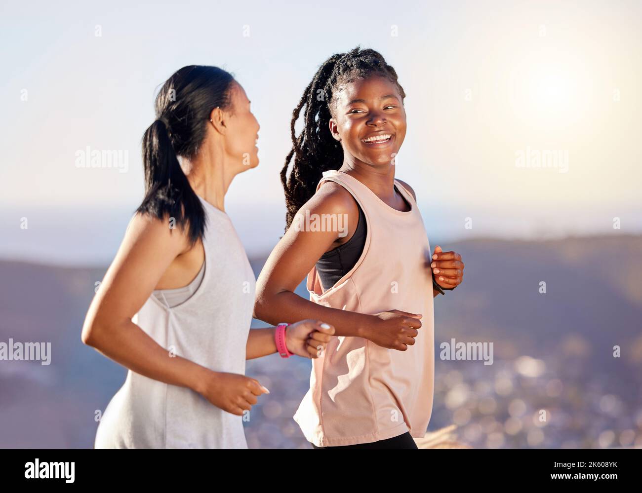 Two happy young female athletes smiling and talking while out for a run on a mountain road on a sunny day. Energetic young women running outdoors to Stock Photo