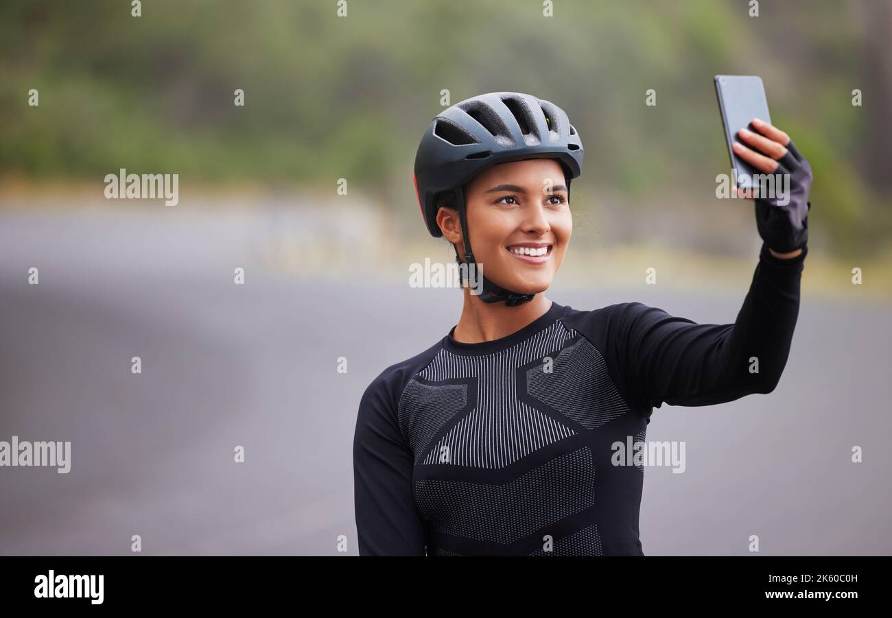 One athletic young woman taking a selfie using her smartphone during a break from cycling outside. Sporty fit mixed race female wearing a helmet and Stock Photo