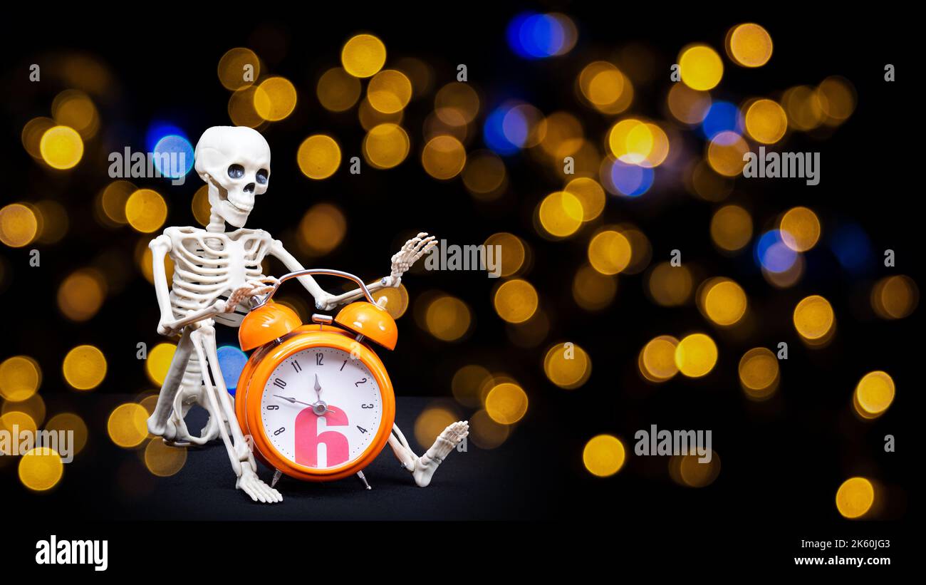 A human skeleton waving his hand, greeting holding an orange alarm clock on a black background, bokeh. It's Halloween time. Greeting card Stock Photo