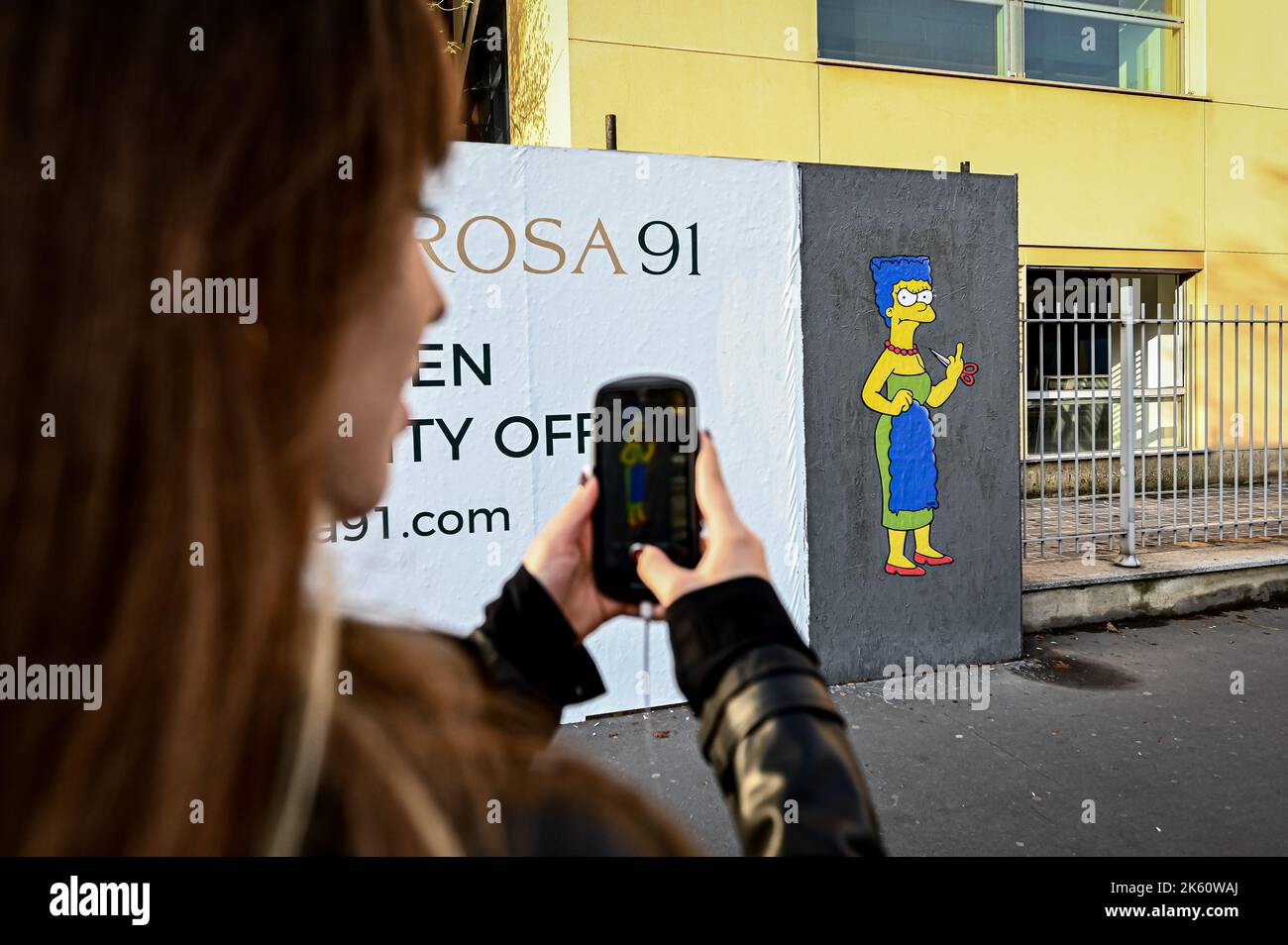 Milan, Italy on October 11, 2022, A woman takes a photo to a mural entitled ‘The Cut 2’ by street artist aleXsandro Palombo depicting The Simpsons' character Marge Simpson cutting her hair and giving the middle finger in solidarity with the women of Iran in front of the Iranian Consulate in Milan, Italy on October 11, 2022. A first version of a similar mural, entitled 'The Cut' appeared on October 5 at the same location and was later on removed. Credit: Piero Cruciatti/Alamy Live News Credit: Piero Cruciatti/Alamy Live News Stock Photo