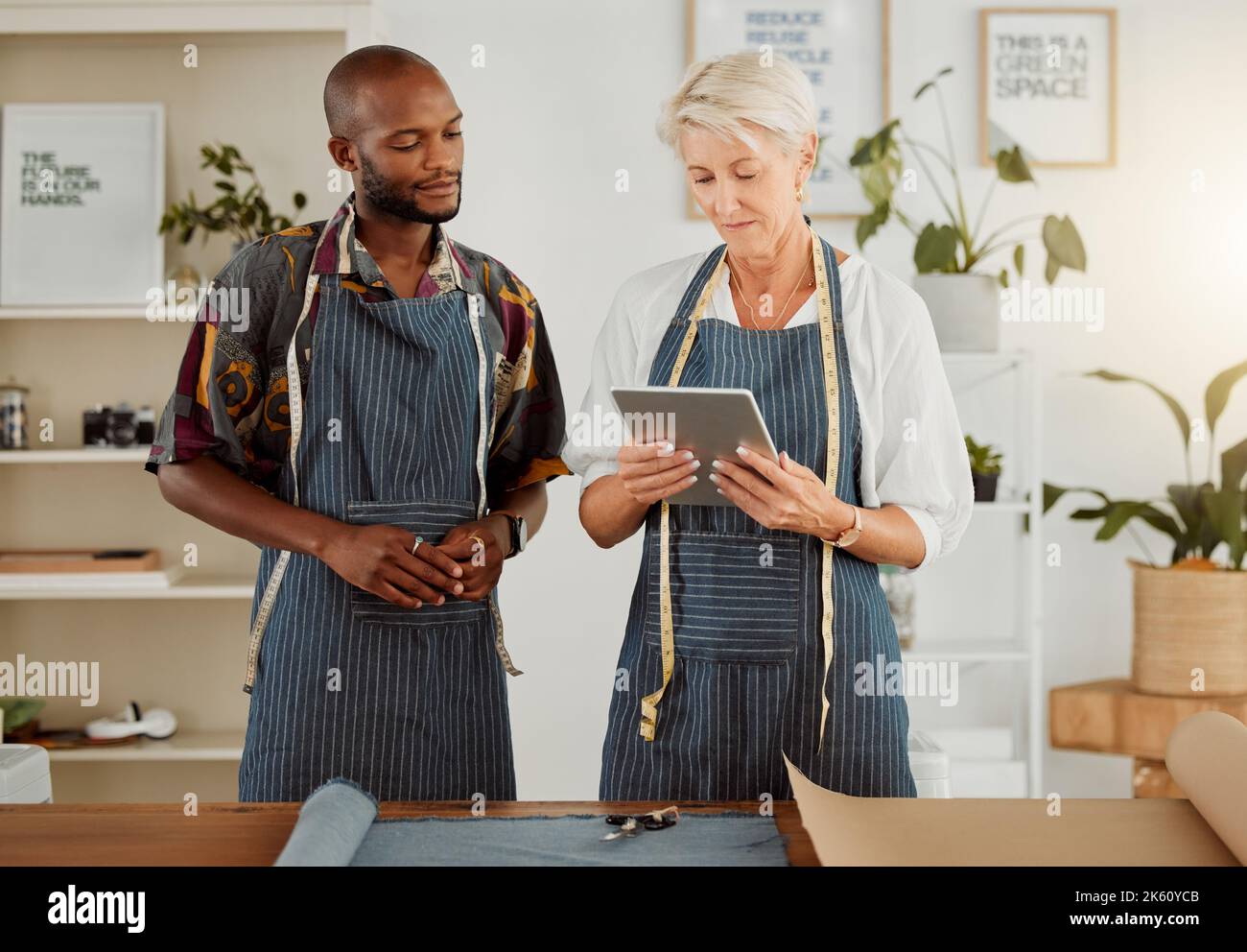 Two designers using a digital tablet while working with material at work. Mature caucasian tailor talking and holding a digital tablet while working Stock Photo