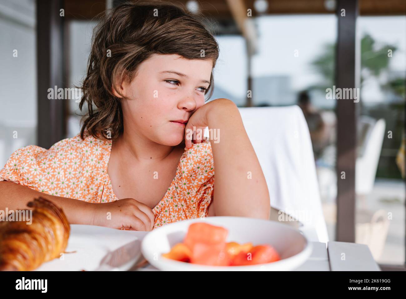 Wistful child with brown hair sitting at table with plates of delicious food in cafeteria and looking away Stock Photo