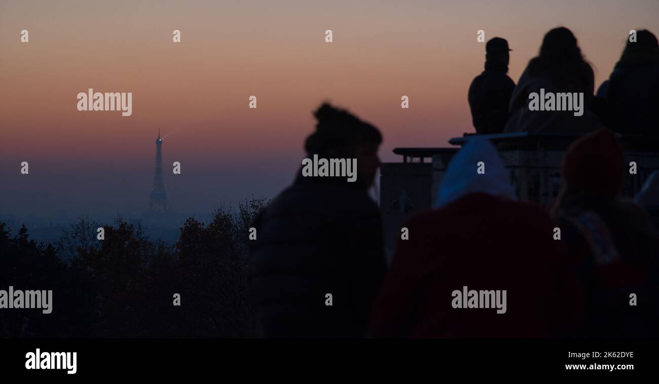 Young people admiring Parisian cityscape view with Eiffel tower at background at sunset and socializing. Belleville park in Paris, France. Stock Photo