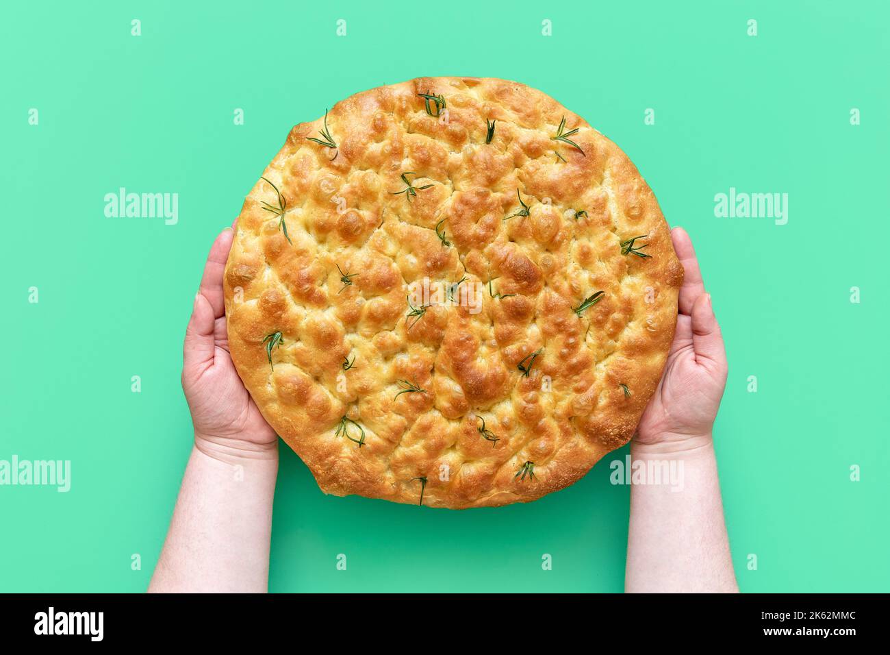 Woman's hands hold a freshly baked rosemary focaccia above a green table. Top view with homemade focaccia with a delicious crust. Stock Photo