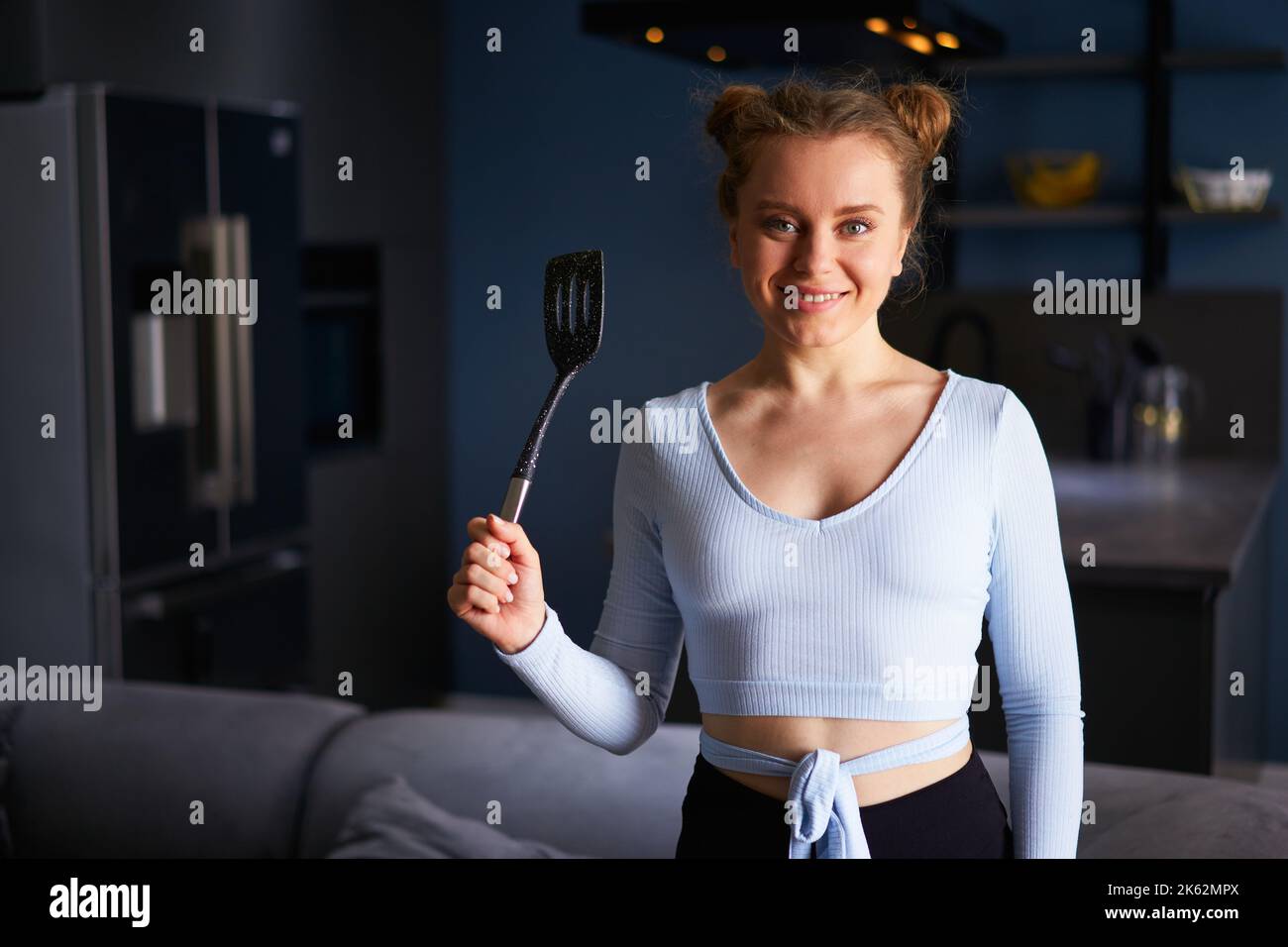 Young caucasian blonde woman holding black spatula smiling happy with teeth standing at home on dark modern stylish kitchen background. Young cheerful Stock Photo
