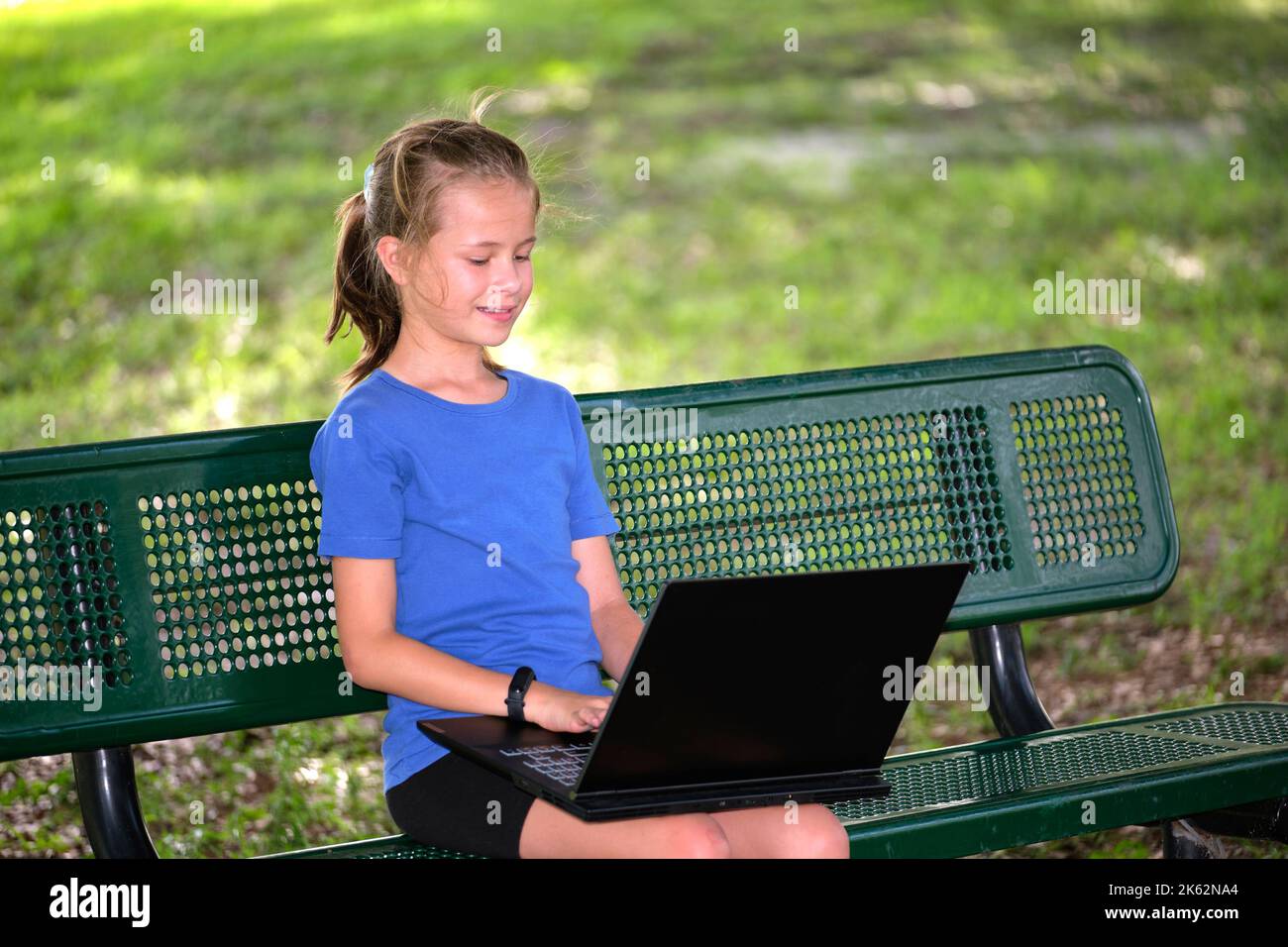 Schoolgirl studying on laptop computer resting on park bench outdoors on summer day. Remote education during quarantine concept Stock Photo