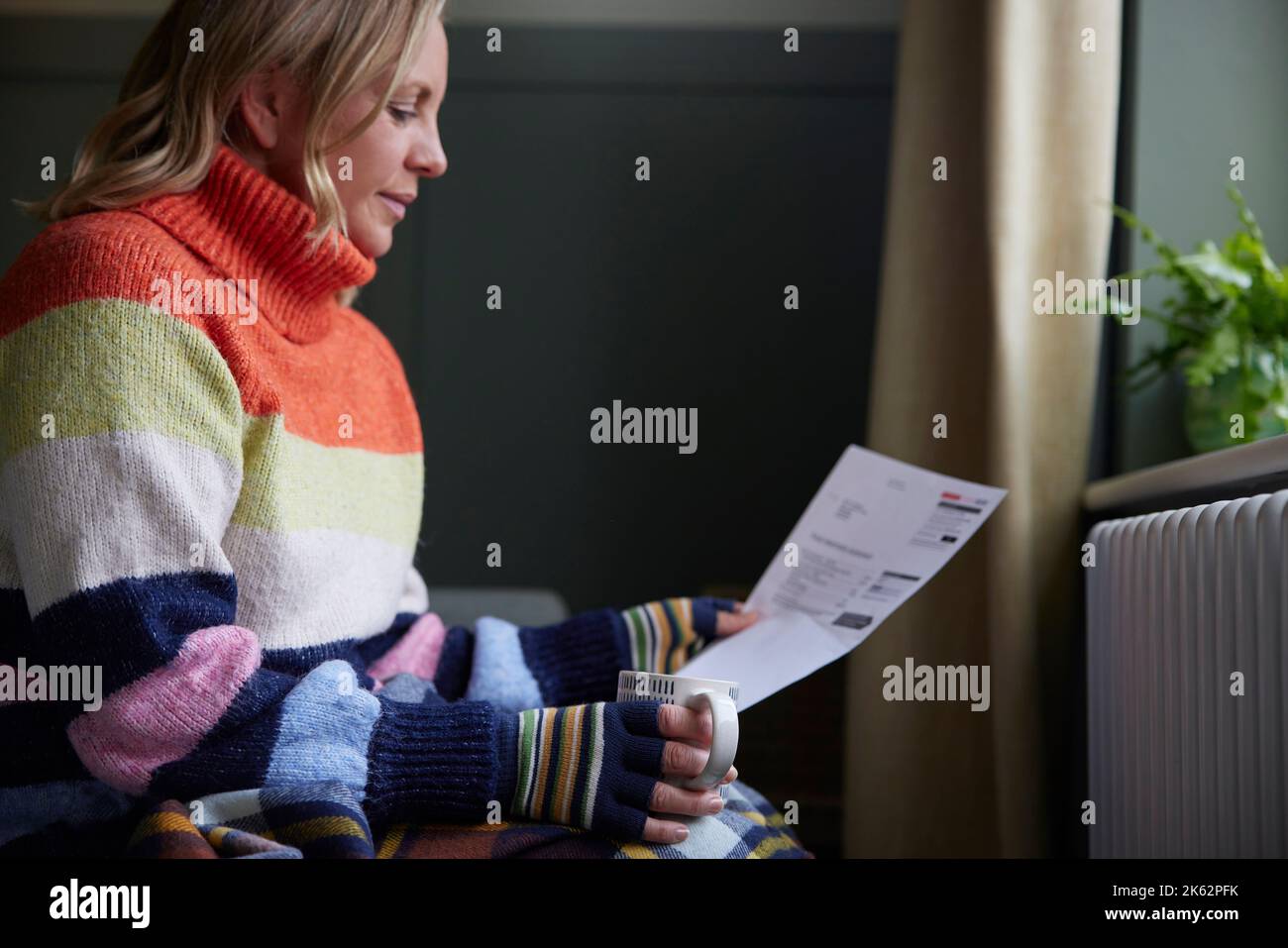 Woman In Gloves With Hot Drink And Bill Trying To Keep Warm By Radiator During Cost Of Living Energy Crisis Stock Photo