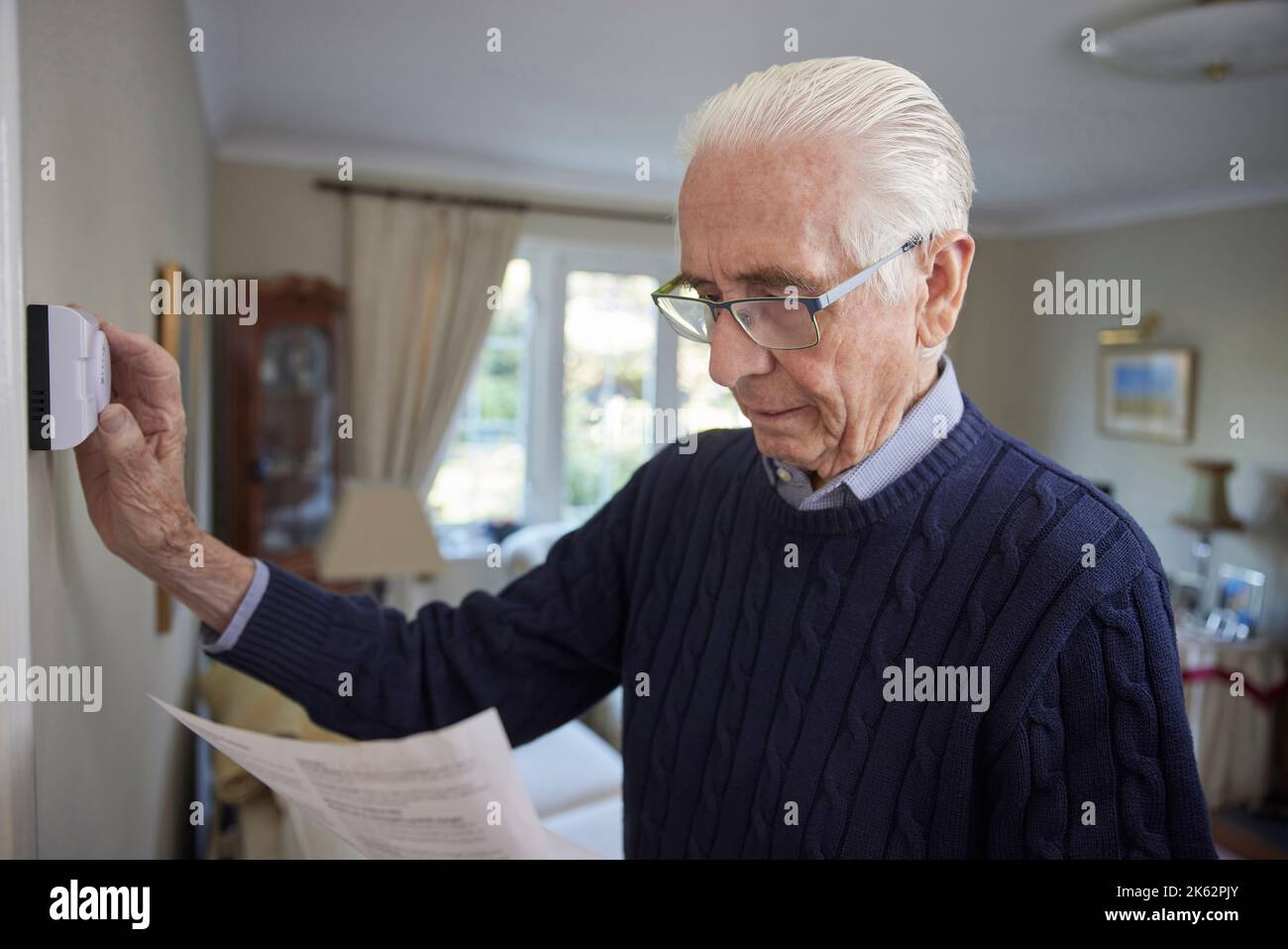 Worried Senior Man With Bill Turning Down Central Heating Thermostat At Home In Energy Crisis Stock Photo