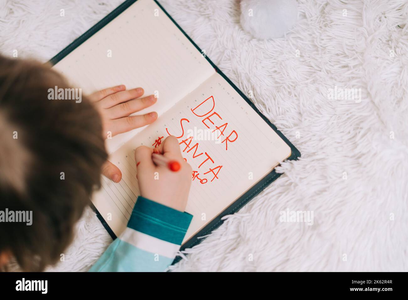 Close up hands of Kid boy, lying on couch and writing letter in note pad to dear Santa at home in Christmas holidays. Child wish list. Dreams of a Stock Photo