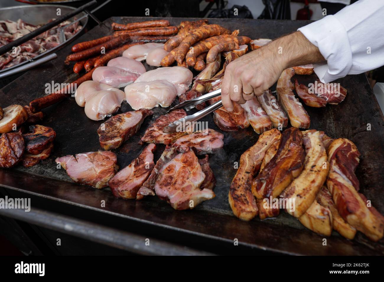 Shallow depth of field (selective focus) details with a man grilling various kinds of meat (pork, chicken, sausages) on a metal hotplate. Stock Photo