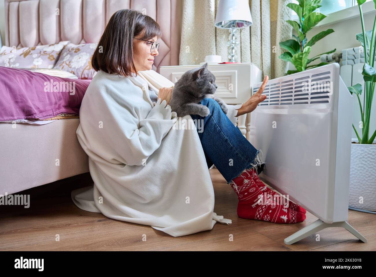 Woman sitting with cat under blanket warming near an electric heater