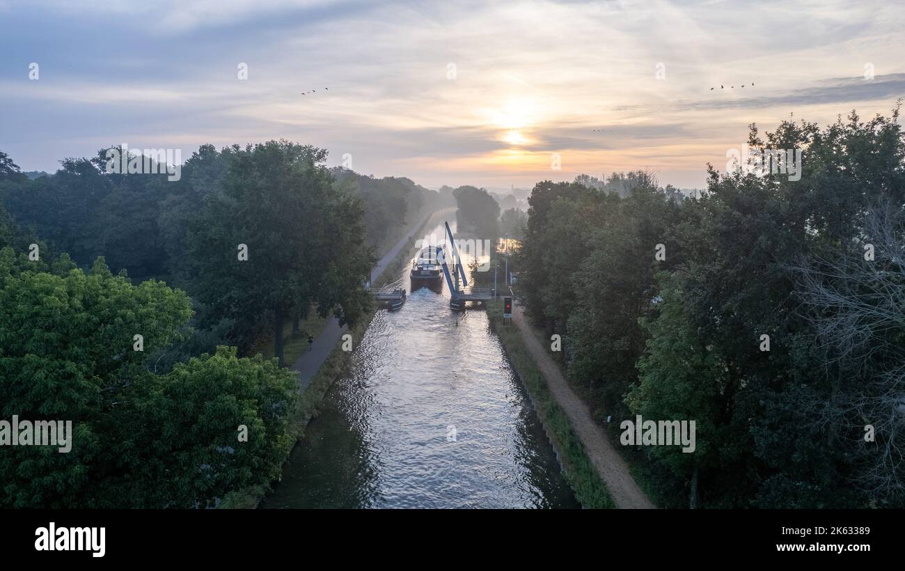 Aerial view of a colourful dramatic sunrise sky over a canal with a cargo boat about to pass under a draw bridge in Belgium. Canals with water for transport, agriculture. Fields and meadows. Landscape aerial view shot from a drone. High quality photo Stock Photo