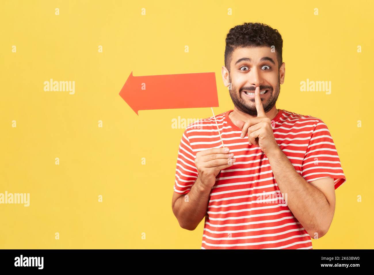 Funny man with beard in T-shirt holding arrow on stick, showing direction and shh gesture holding finger near lips, with naughty smile on face. Indoor studio shot isolated on yellow background. Stock Photo