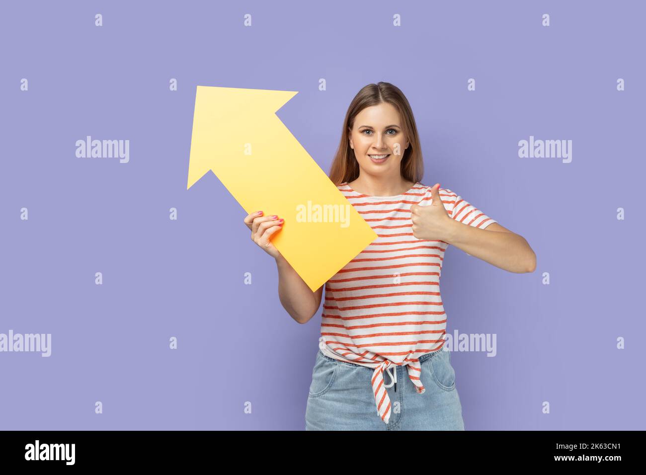 Portrait of pretty smiling blond woman wearing striped T-shirt holding big yellow arrow pointing aside and showing thumb up, like gesture. Indoor studio shot isolated on purple background. Stock Photo