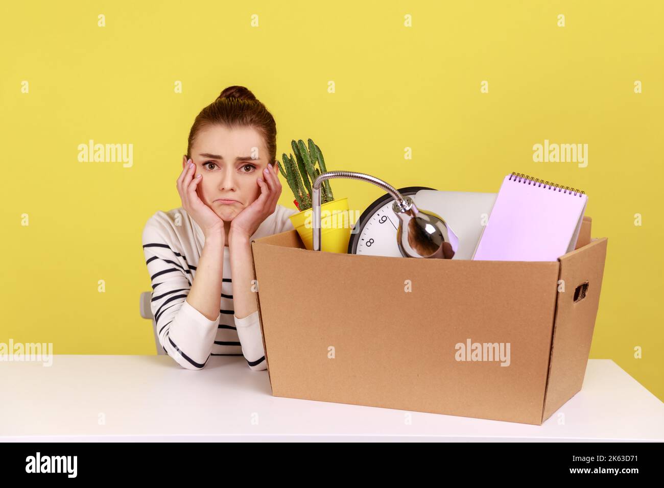 Unhappy depressed woman office worker sitting at workplace with cardboard box with her things, dismissal, bankruptcy, keeps hands under chin. Indoor studio studio shot isolated on yellow background. Stock Photo