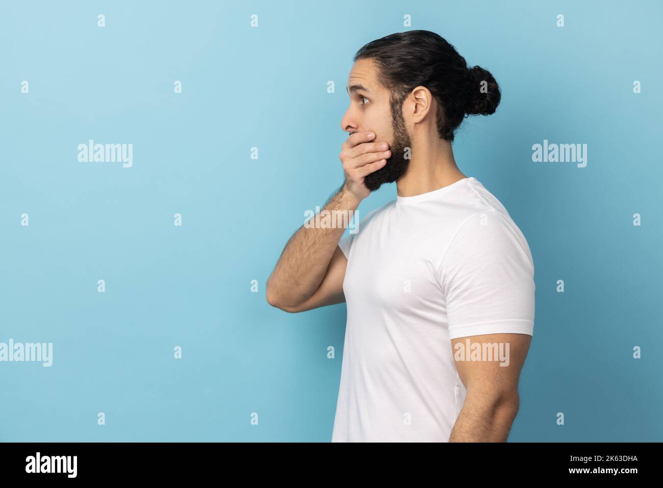 Side view of man with beard wearing white T-shirt covering mouth, doesnt want to spread rumours, shocked to hear sudden news from interlocutor. Indoor studio shot isolated on blue background. Stock Photo