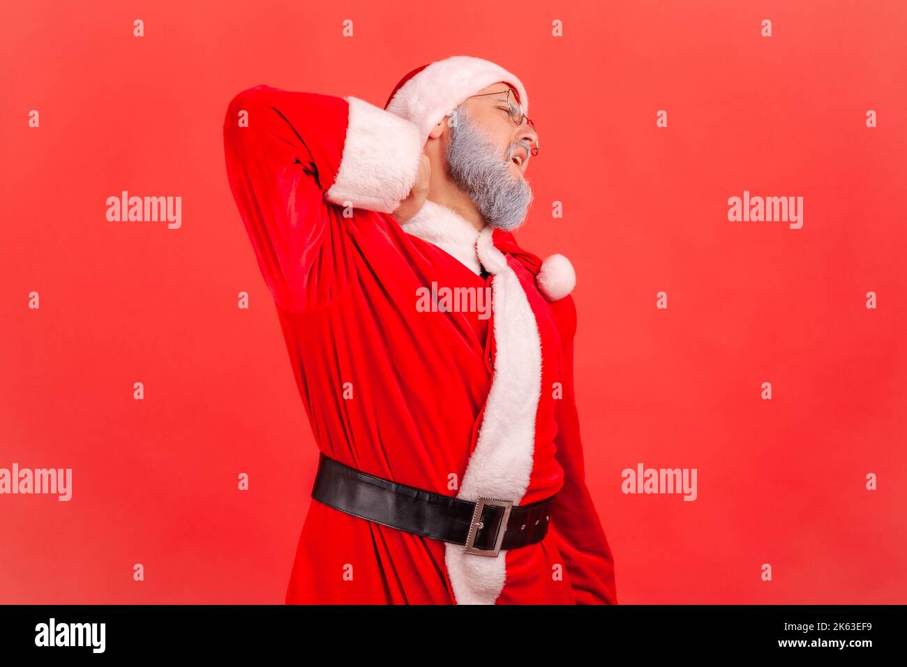Shoulders and neck pain. Portrait of elderly man with gray beard wearing santa claus costume massaging neck to relieve pain, muscle strain in back. Indoor studio shot isolated on red background. Stock Photo