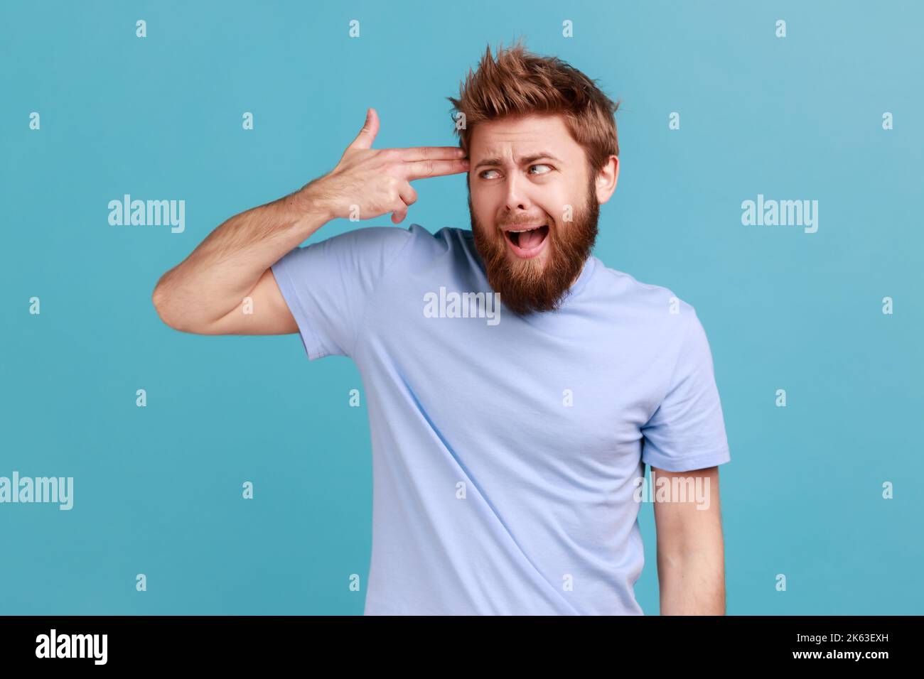 Depression, suicide gesture. Portrait of crazy bearded man standing with finger gun pointed to head, shooting himself with hand pistol. Indoor studio shot isolated on blue background. Stock Photo