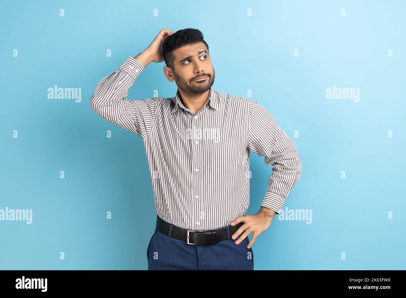 Portrait of young pensive businessman with beard touching head and thinking about important question, making hard decision, wearing striped shirt. Indoor studio shot isolated on blue background. Stock Photo